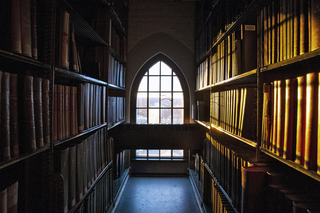 the setting sun spills through an arched window into a dark aisle, illuminating the library stacks